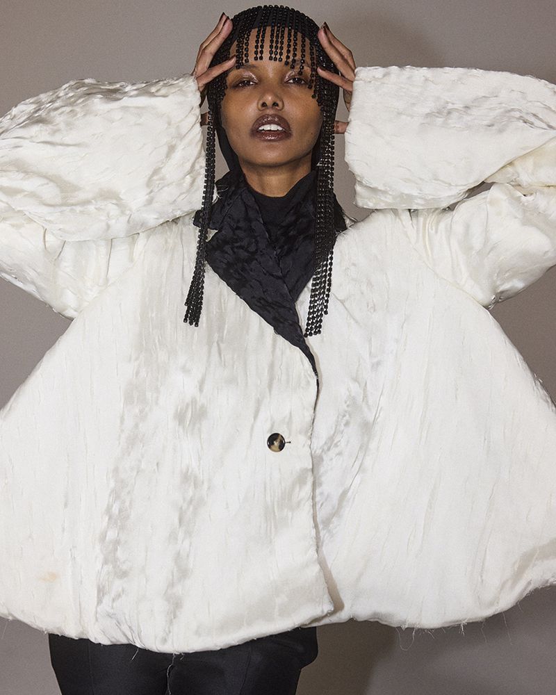 Woman wearing a white, short, puffy jacket with a black scarf and beaded fringe headpiece in a studio environment.