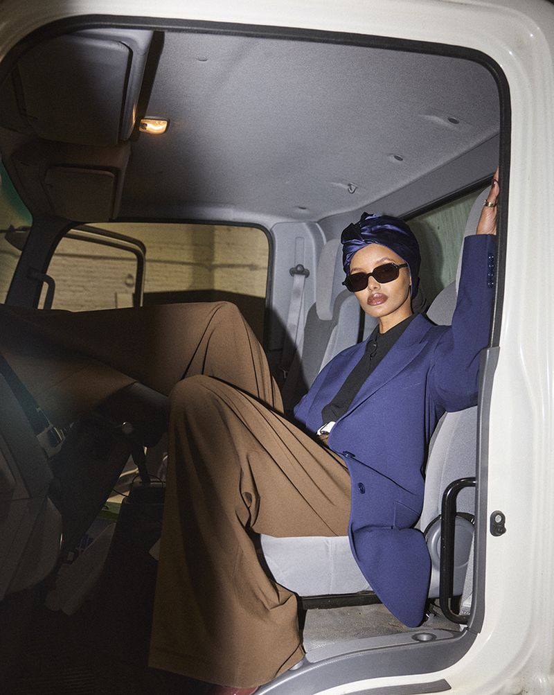 Woman wearing a blue blazer and brown pants sitting inside of a white truck cab, posing with sunglasses and a head wrap.