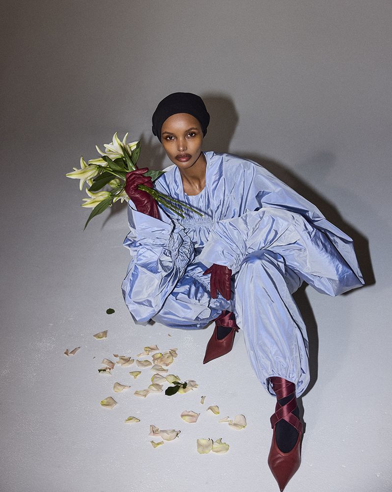 Fashion shot of a woman in a light blue outfit and maroon shoes, squatting and holding a bouquet of lilies in an indoor studio.