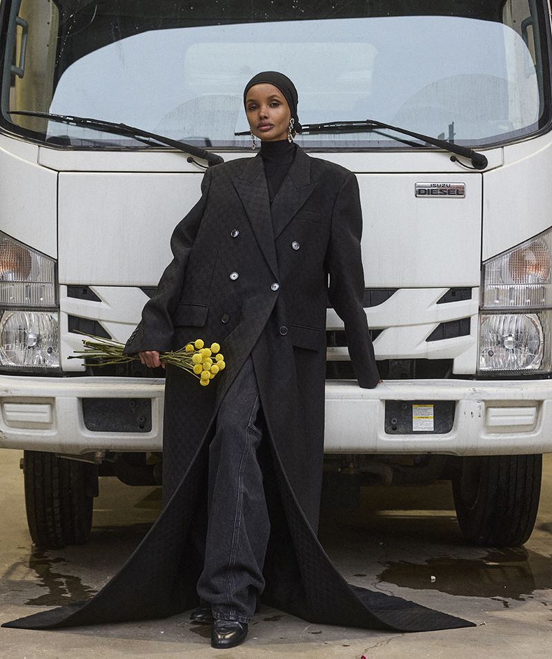 Adult female in black coat and headscarf poses in front of white truck holding yellow flower bouquet in fashion style editorial.