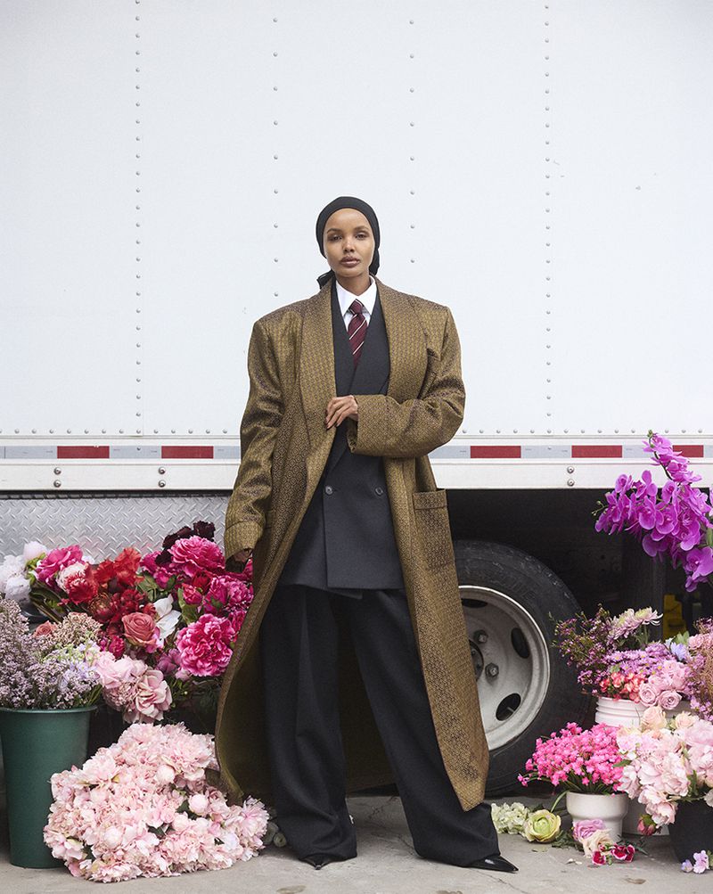 A person wearing a dark suit, a patterned brown overcoat, and a headscarf, stands among a floral display in front of a white truck.