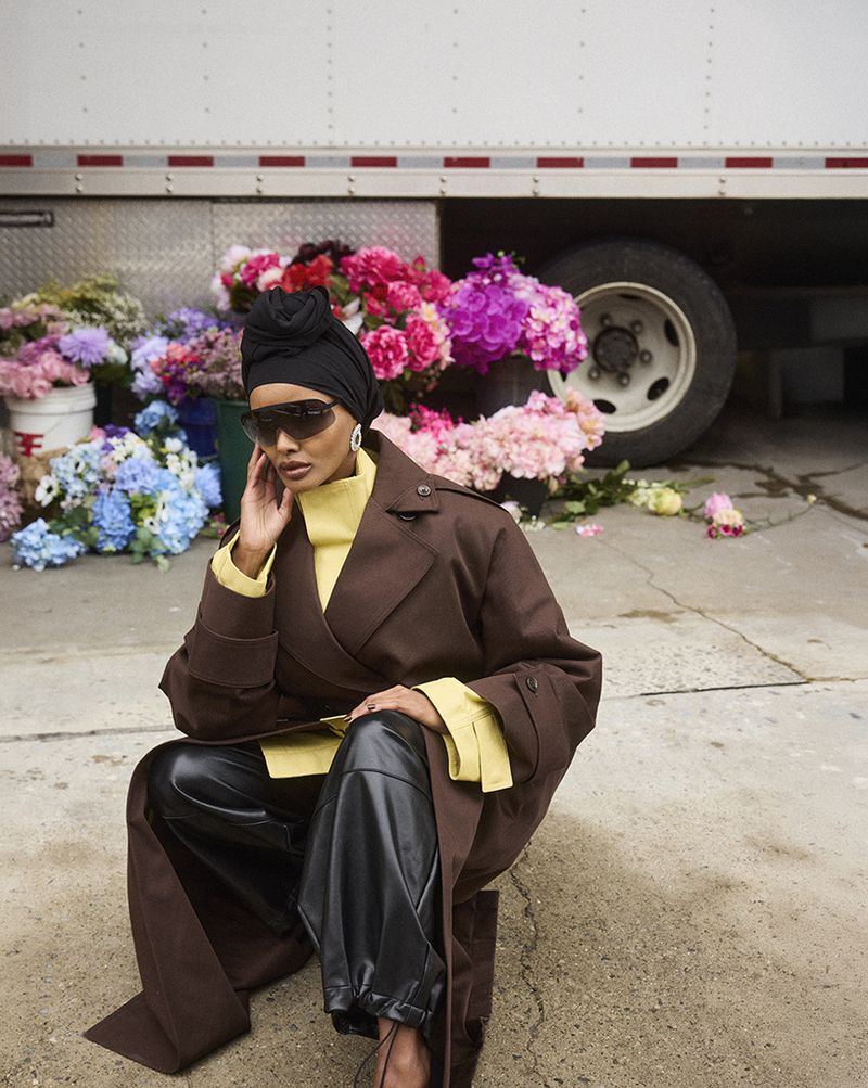 An adult female poses in a brown coat and black pants against a backdrop of colorful flowers and a white truck, wearing sunglasses and a head covering.