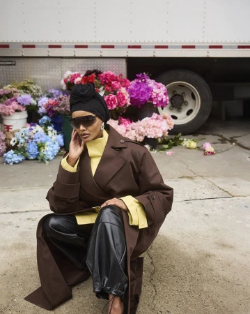 An adult female poses in a brown coat and black pants against a backdrop of colorful flowers and a white truck, wearing sunglasses and a head covering.