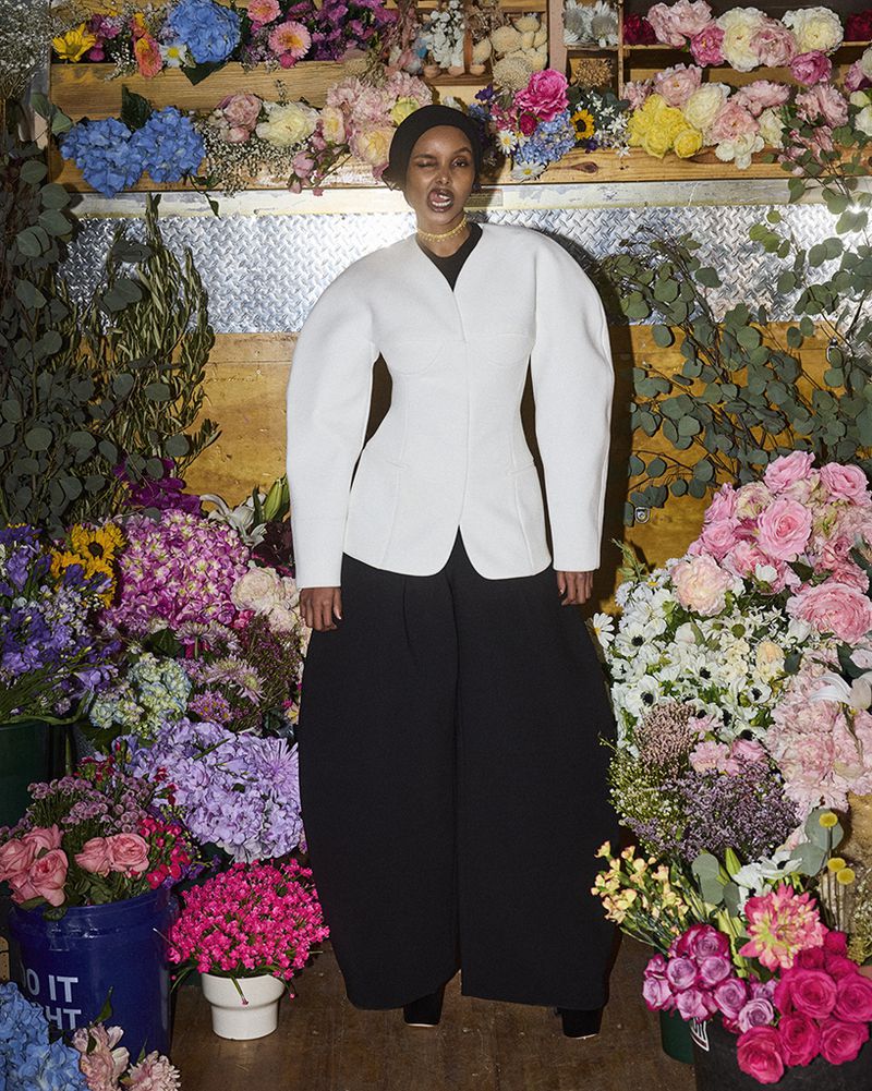 A woman in a white jacket and black pants poses among colorful flowers, creating a striking fashion portrait with a vibrant, natural backdrop.