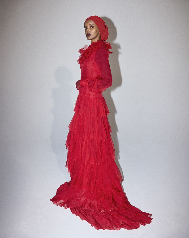 Elegant adult female fashion model in a full-length red gown and matching head covering, standing in a photo studio with a white backdrop.