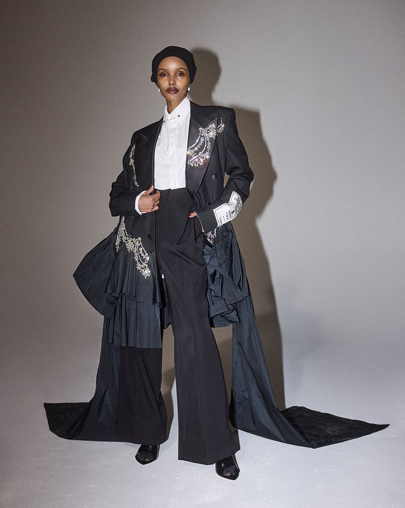 An adult female poses in a photo studio wearing a black jacket with silver details, a white collared shirt, and black pants. 
