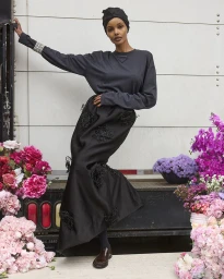 Halima Aden in dark long-sleeve top and black embellished skirt surrounded by colourful floral arrangements for Vogue Arabia.