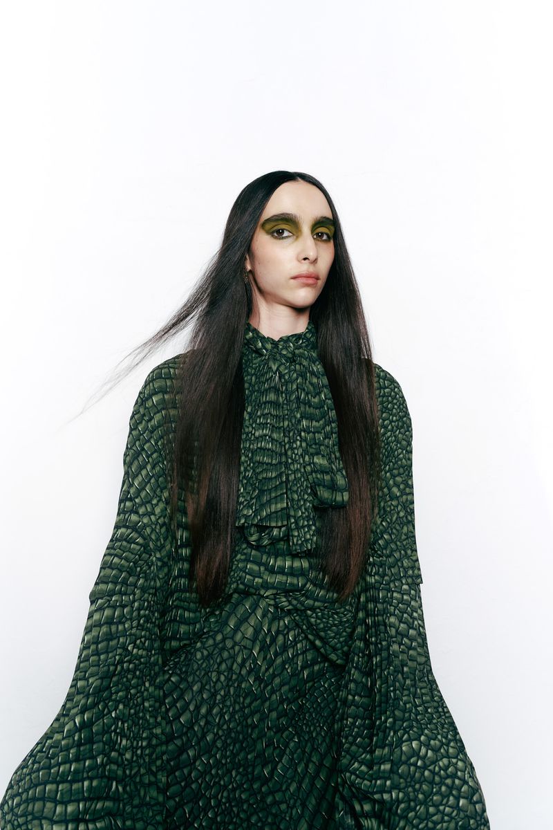 Woman with long dark hair and green eye makeup poses in a full-length dress with a green and black alligator scale pattern against white.