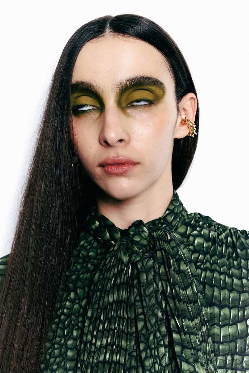 A fashion portrait of an adult woman with olive green eye makeup and a high-neck green dress with a scale pattern on a white background.