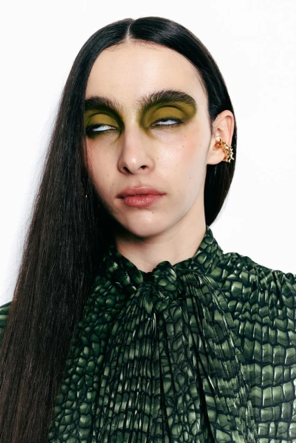 A fashion portrait of an adult woman with olive green eye makeup and a high-neck green dress with a scale pattern on a white background.