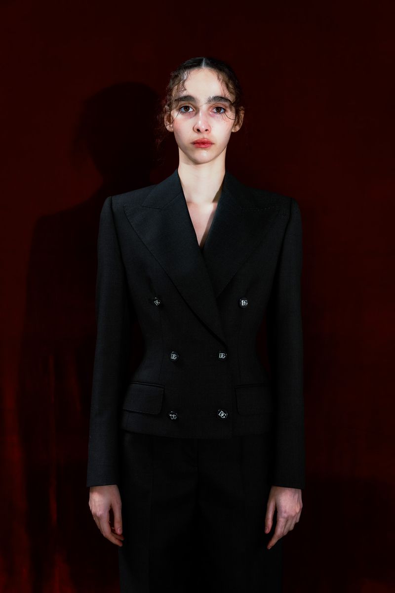 Adult female poses in a formal black double-breasted blazer and skirt against a dark red background in a studio setting.