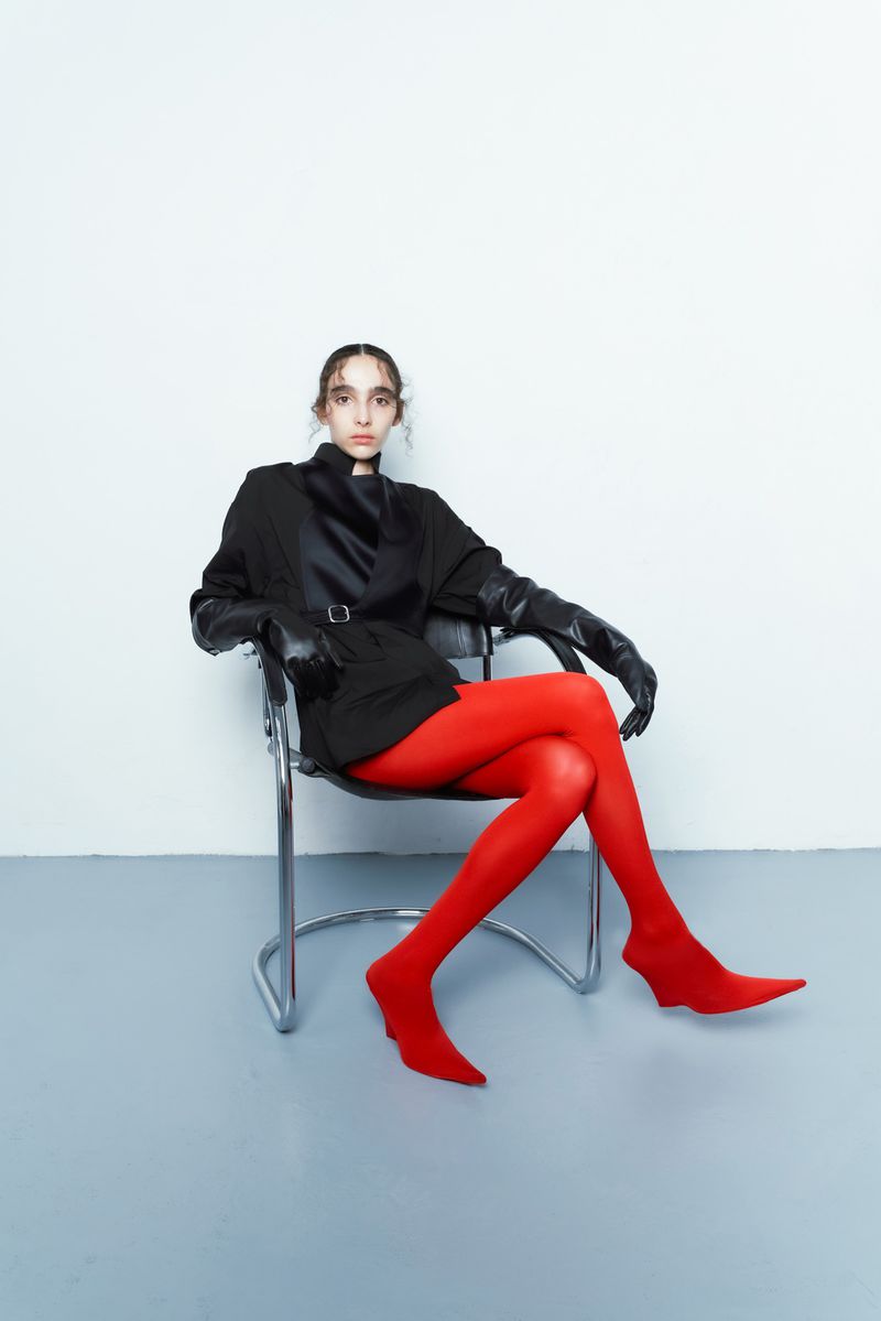 A woman poses in a black jacket, gloves, and red stockings, seated in a chrome chair against a white wall in a studio setting.
