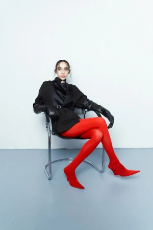 A woman poses in a black jacket, gloves, and red stockings, seated in a chrome chair against a white wall in a studio setting.