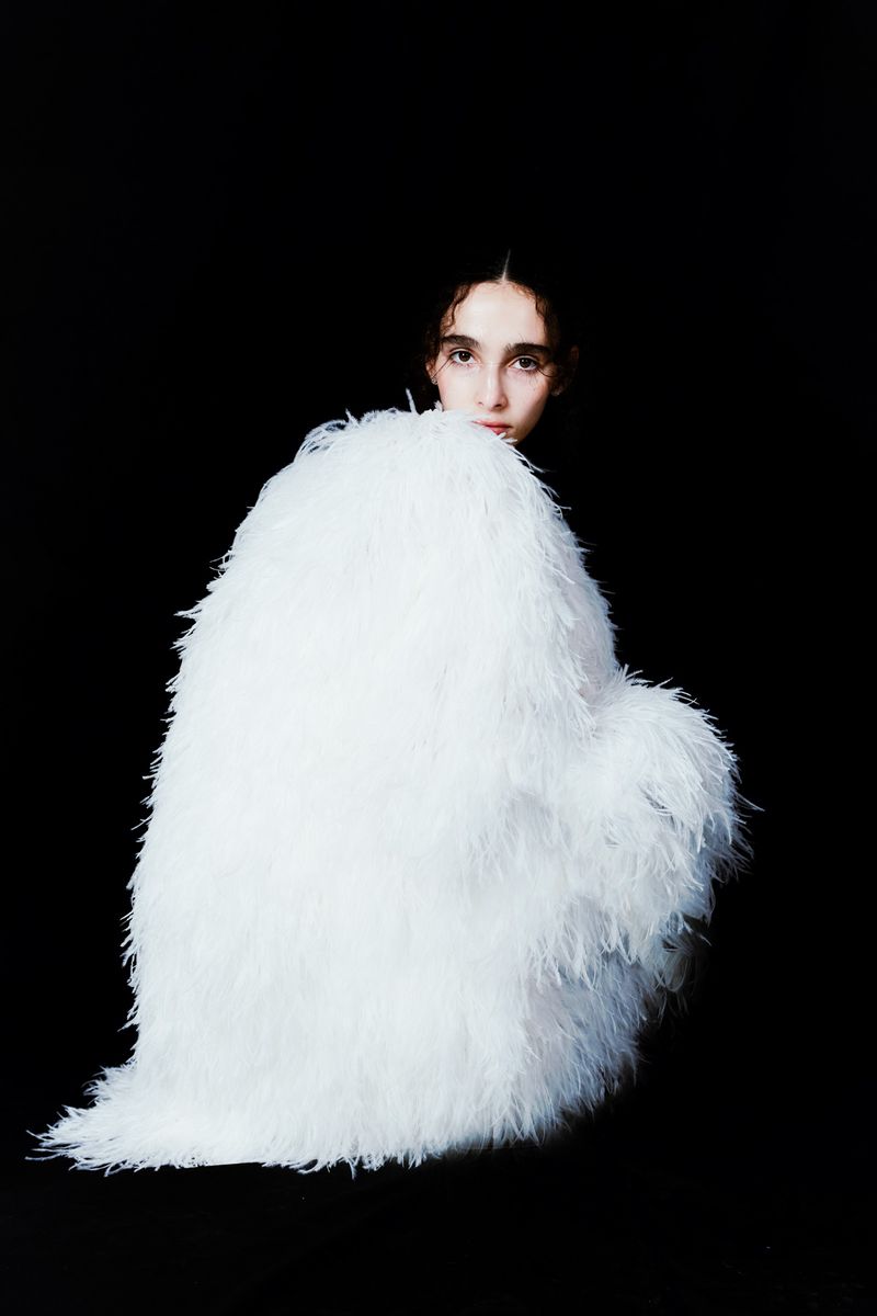 Fashion shot of a woman partially obscured by a large, furry white jacket against a black background, with minimal post-production.