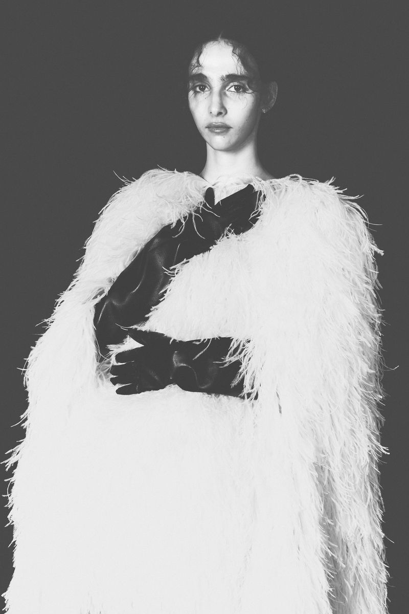 Monochrome portrait of a woman in a feathered wrap and gloves against a dark backdrop, emphasizing texture and minimalist elegance.