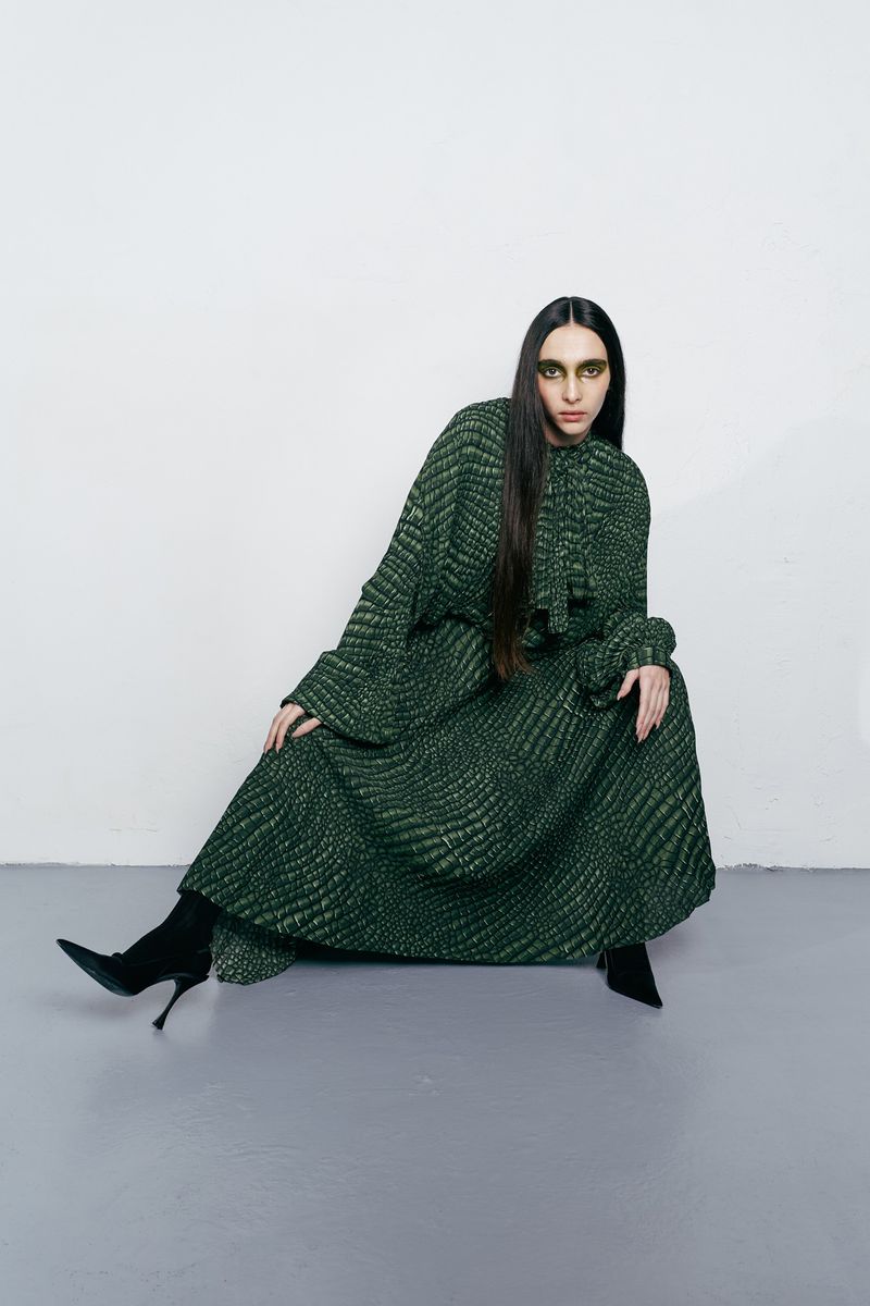 A woman wearing a long, patterned green dress and black boots sits in a photo studio with a light grey floor and white background.