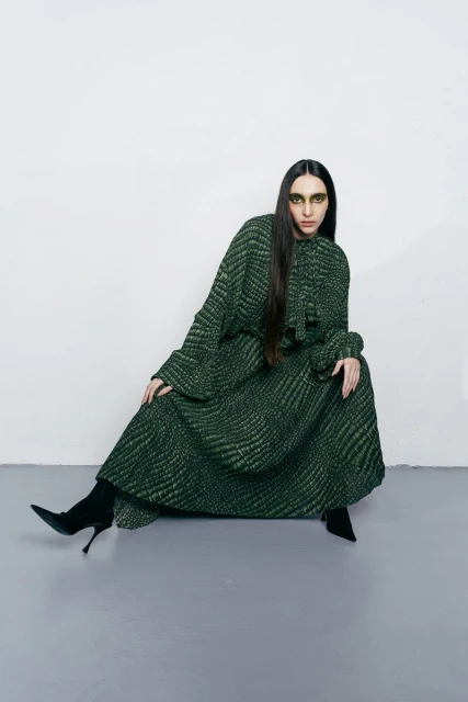 A woman wearing a long, patterned green dress and black boots sits in a photo studio with a light grey floor and white background.