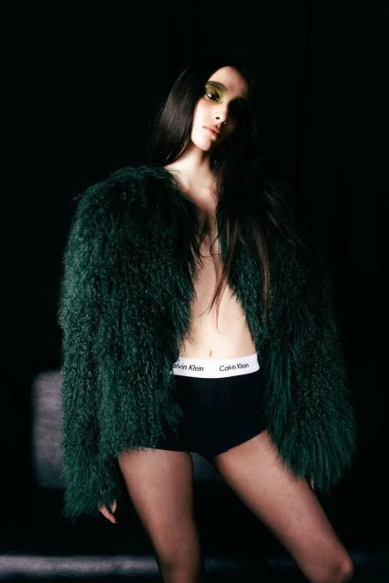 A woman poses indoors wearing a shaggy, green jacket and Calvin Klein underwear, with bright eyeshadow in a dark, moody setting.