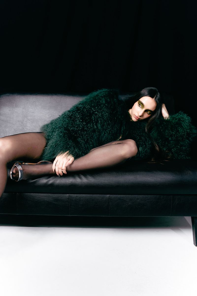 A woman in a green textured coat and black tights reclines on a dark leather couch against a black and white background.