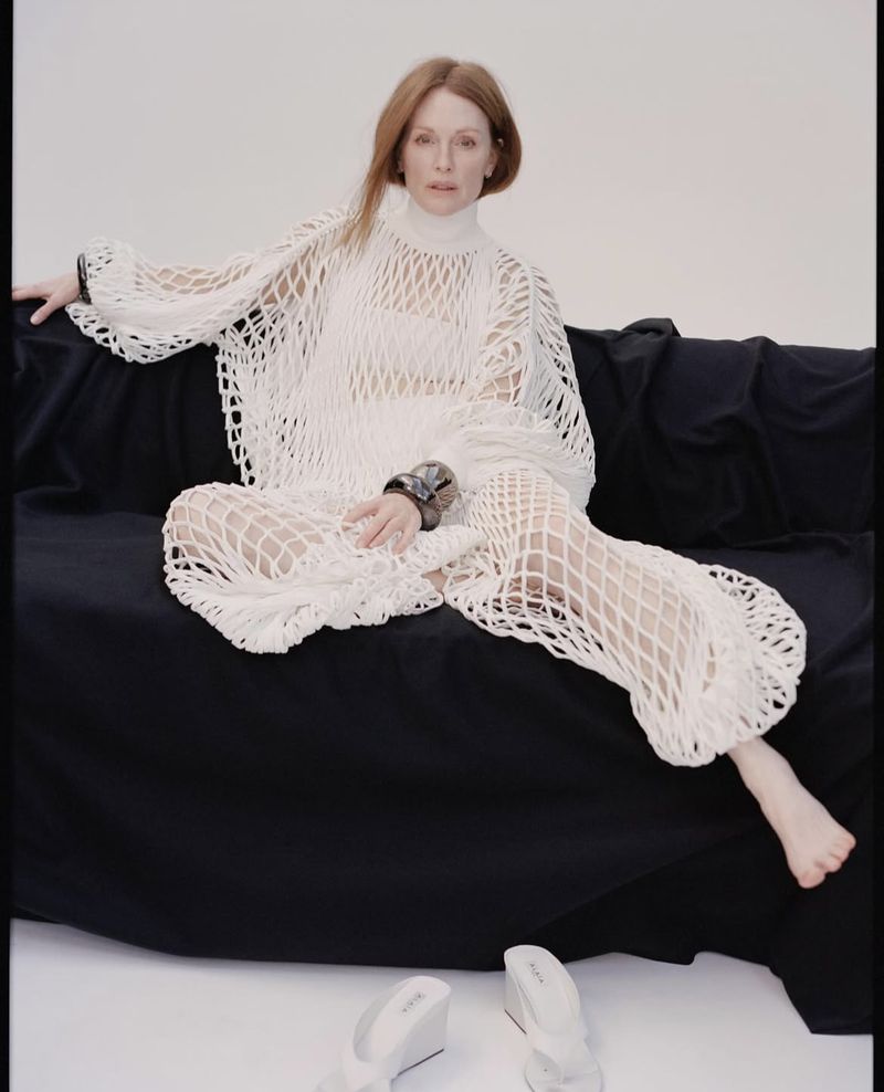 Elegant red-haired woman sits on a black couch in a white net dress with shoes on the floor, set against a bright white backdrop.