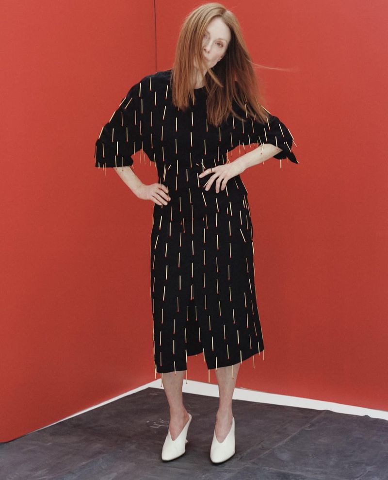 A fair-skinned woman with red hair poses in a black midi dress with gold embellishments against a solid red background.