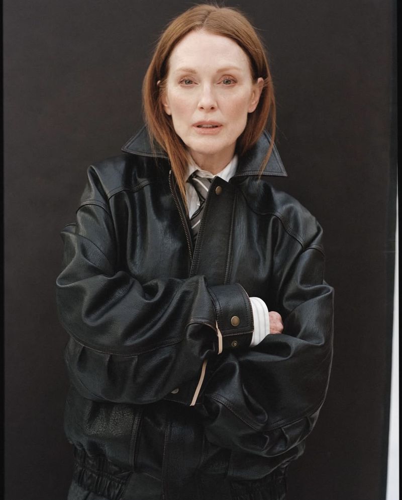 Headshot of a red-haired adult female wearing a black leather bomber jacket and striped tie against a dark, neutral background.