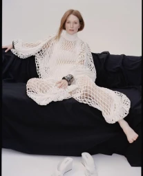 Julianne Moore seated on a dark fabric sofa wearing a white open-knit garment against a neutral background