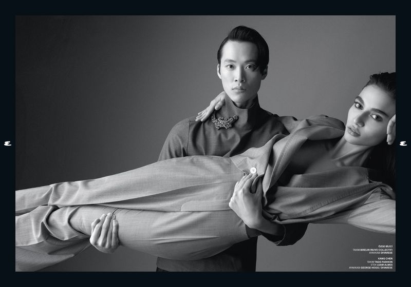 Monochrome fashion shot features an adult male carrying an adult female, both wearing grey tailored suits, against a grey backdrop.