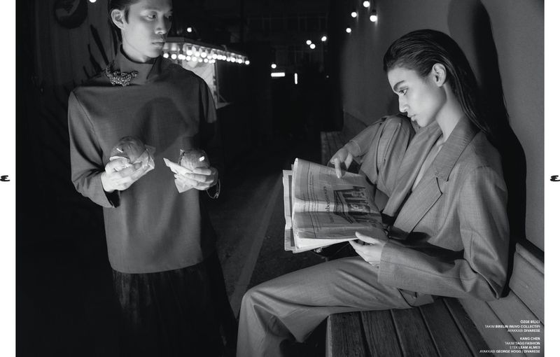 Monochrome fashion shot features two models: a man in a dark top holding food, and a woman in a suit reading a newspaper.