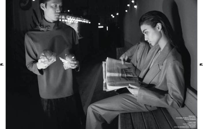 Monochrome fashion shot features two models: a man in a dark top holding food, and a woman in a suit reading a newspaper.