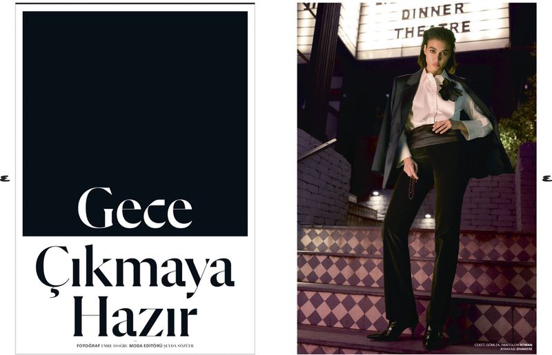 Woman in tailored black suit stands on patterned stairs in front of a theater at night, fashion editorial, stylish evening wear.