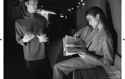 Monochrome fashion editorial image showing a standing man holding wrapped food and a seated woman reading a newspaper