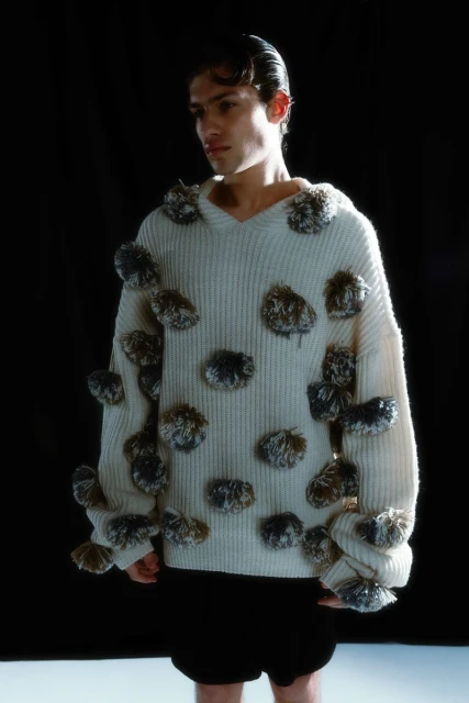A young adult man in a cream-colored sweater with decorative pompoms, standing indoors against a dark background in a fashion setting.