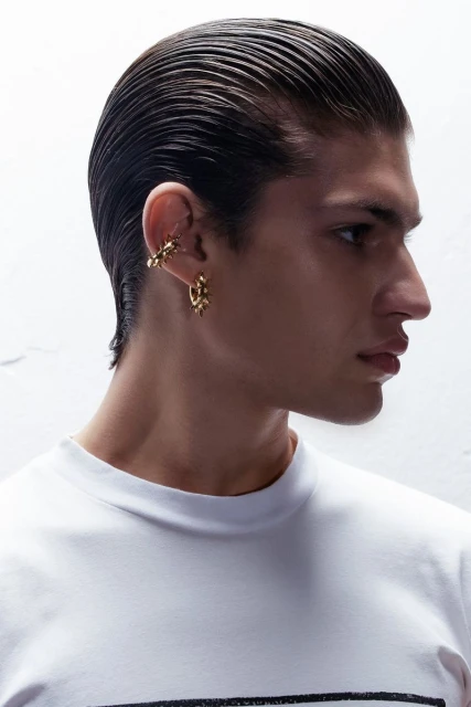 Profile shot of a young adult male with slicked-back hair wearing gold jewelry and a white t-shirt against a textured white background.