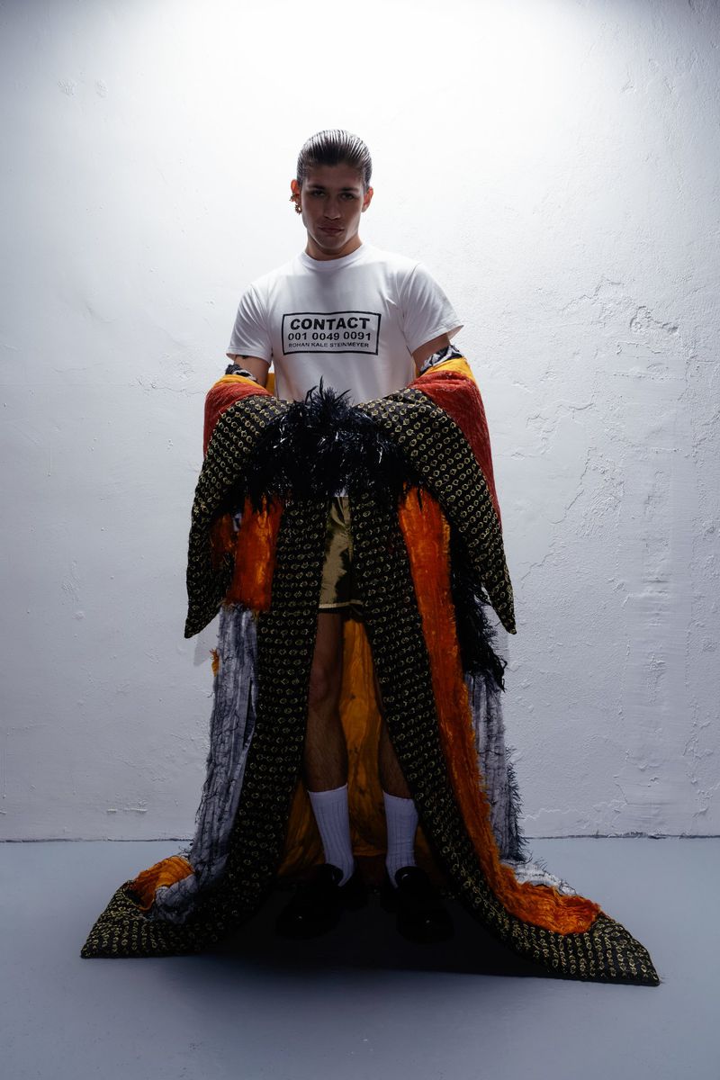 Male model in studio wearing a white t-shirt, patterned coat, and white socks, posing against a simple white wall with overhead lighting.