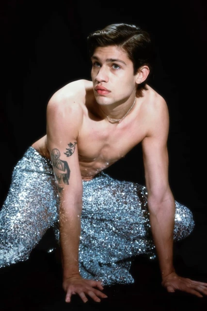 Young adult male in a crouching pose wearing silver sequin pants against a dark background in a studio portrait with tattoo on arm.