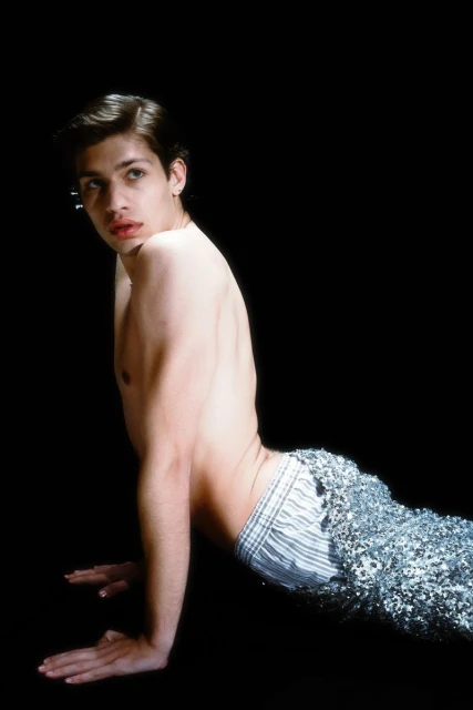 Shirtless male model with dark hair poses in a sequin-covered fishtail against a black backdrop, showcasing beauty and fashion elements.