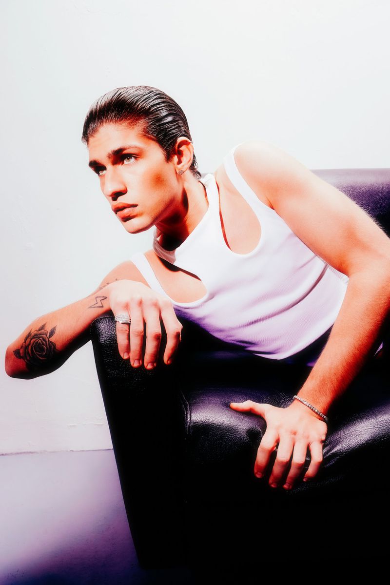 Young man in white tank top poses in black chair against white wall, captured in studio lighting with slicked-back hair. Fashion shot.