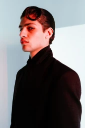 Portrait of a male model wearing a high-collared black jacket with styled wet-look hair against a white and light blue wall.