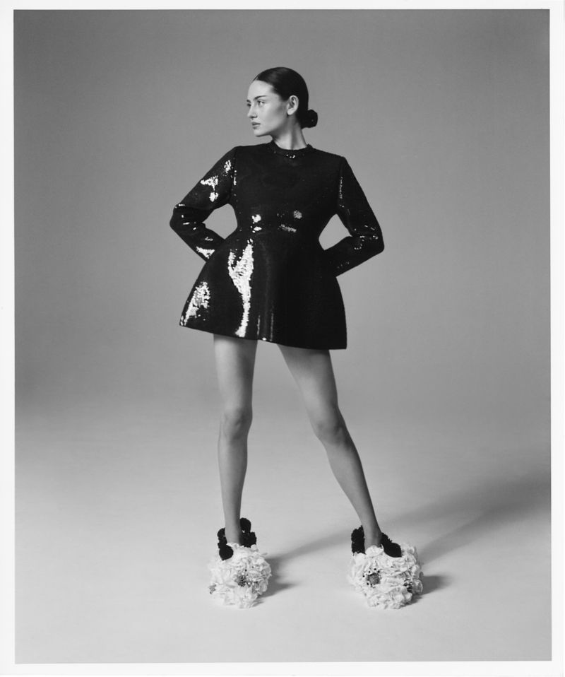 Monochrome fashion portrait of a woman in a shiny, short black dress and sculptural floral shoes against a plain grey backdrop.