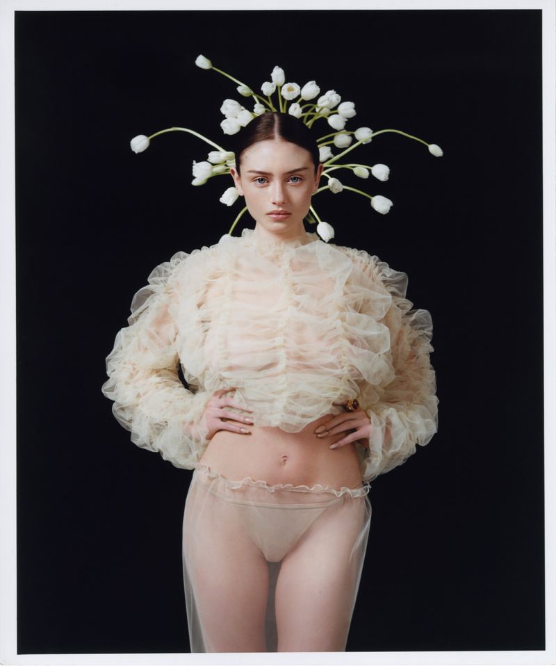Portrait of a young woman wearing a sheer ruffle top, tulle skirt, and floral headpiece against a solid black backdrop in a studio setting.