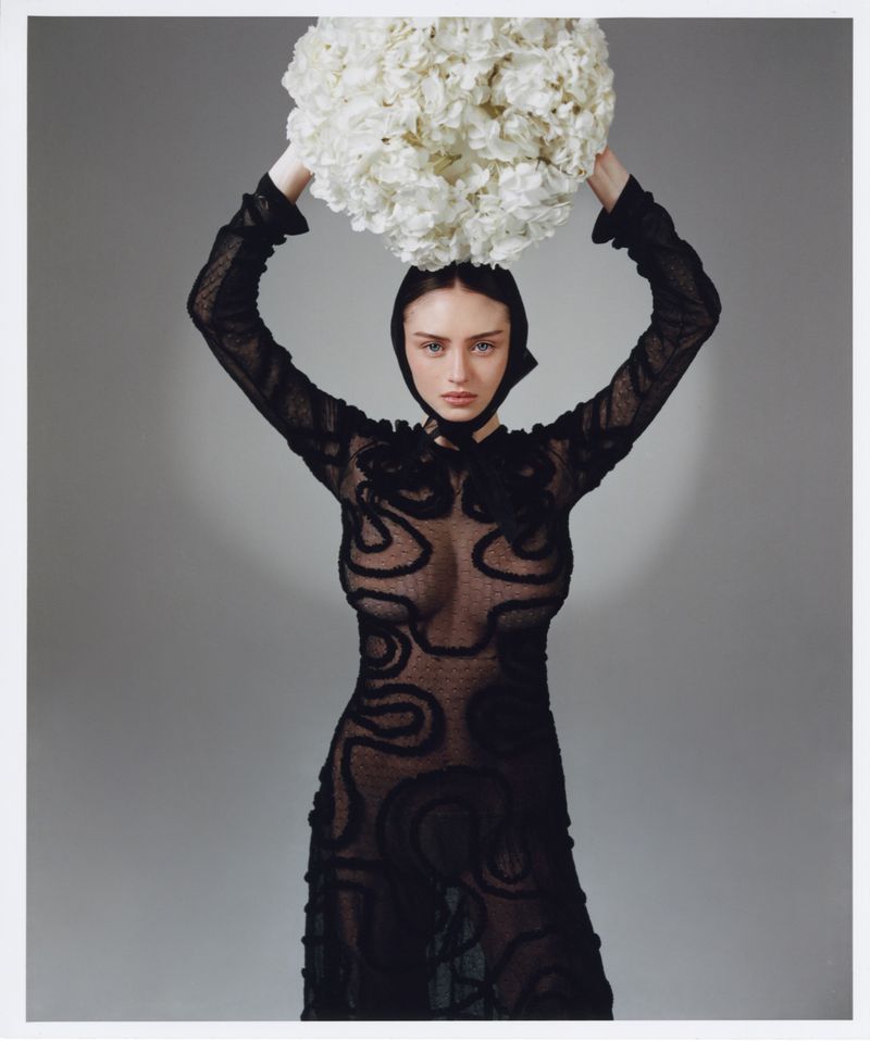 A woman in a patterned black dress stands in front of a gray backdrop, holding a ball of white flowers above her head in a studio shot.