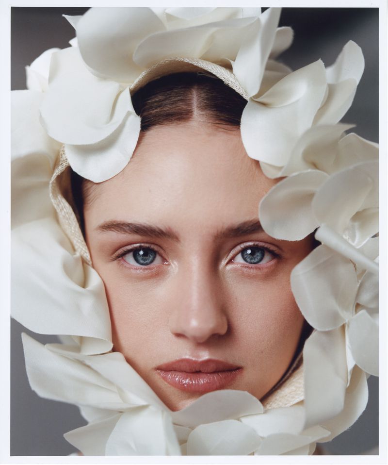 Close-up portrait of a woman with light skin, blue eyes, and neutral expression, framed by white floral headpiece in soft studio lighting.