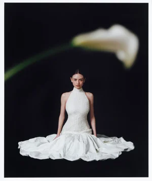 Model Leni Klum in a white halterneck dress seated against a black background with a blurred flower in the foreground.