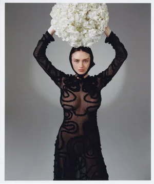 Model Leni Klum holding a large white floral arrangement above her head in a black dress against a grey seamless background