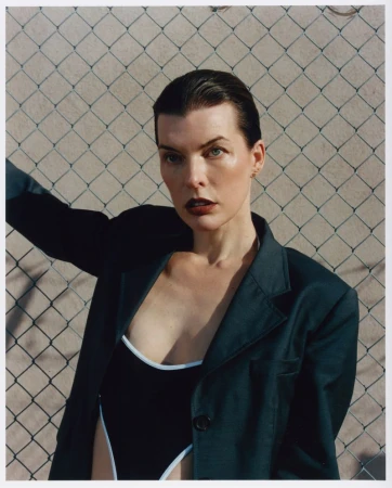 Milla Jovovich poses in a black swimsuit and dark blazer against a chain-link fence in a medium shot, capturing a minimalist fashion aesthetic.