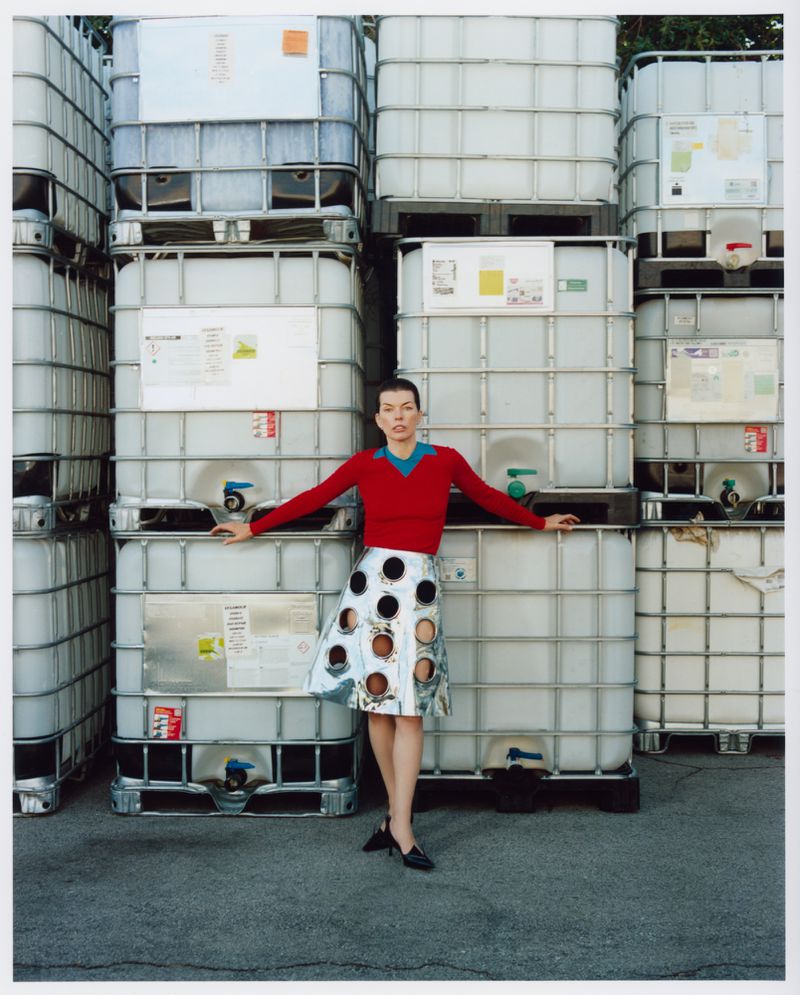 A woman poses in front of stacked intermediate bulk containers (IBCs) wearing a red sweater and patterned skirt, with black shoes.
