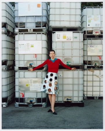 A woman poses in front of stacked intermediate bulk containers (IBCs) wearing a red sweater and patterned skirt, with black shoes.