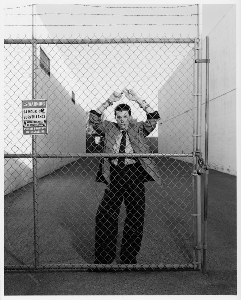 A man in a suit stands behind a chain-link fence in an alleyway, hands raised above his head. A surveillance sign is visible on the fence.