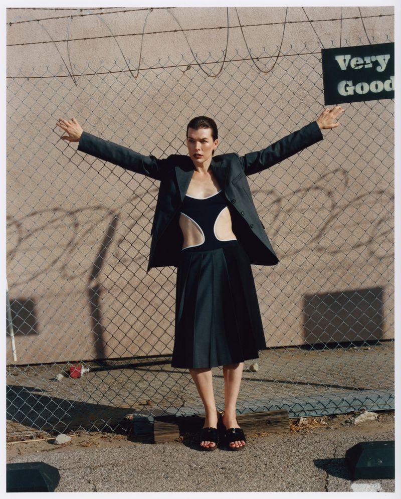 Adult woman in a grey blazer and black skirt stands in front of a chain link fence with a sign that reads 'Very Good'.